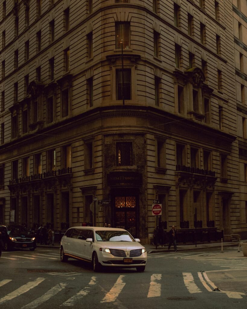 A stylish limousine navigates a bustling city street corner surrounded by towering architecture.